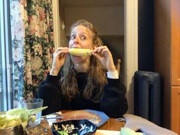 A woman with long hair, wearing a black sweater, sits at a table and enthusiastically eats corn on the cob, surrounded by plates, utensils, and floral curtains.