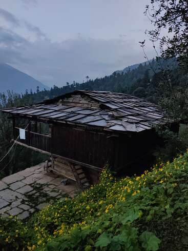 Das Bild zeigt ein rustikales Holzhaus mit Ziegeldach, eingebettet zwischen Bäumen und Blumen, vor einer Bergkulisse unter einem wolkenverhangenen Himmel.