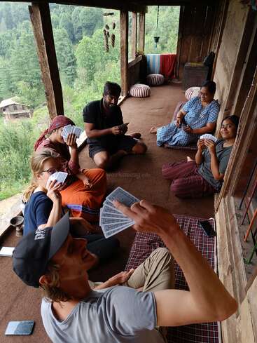Das Bild zeigt eine Gruppe von sechs Personen, die auf einer Veranda mit Blick auf Bäume und ein Haus im Hintergrund Karten spielen und alle Karten in den Händen halten.