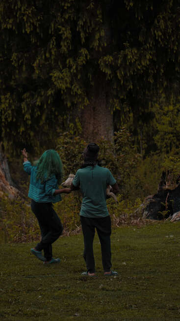 Das Bild zeigt zwei Personen, die auf einer Wiese stehen, wobei eine Person eine Tasche hält und die andere auf einen Baum im Hintergrund deutet.