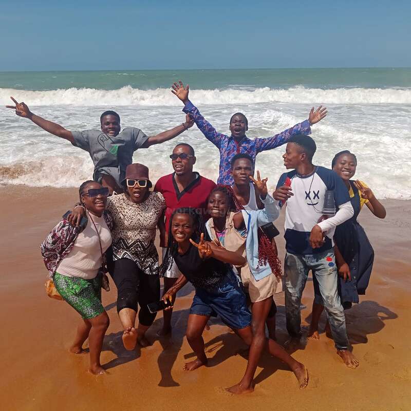Un alegre grupo de amigos posa en una playa soleada con las olas rompiendo a sus espaldas, sonriendo, riendo y haciendo signos de paz, disfrutando claramente del momento juntos.