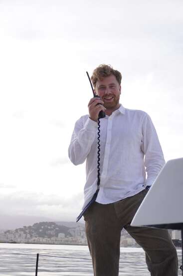A imagem mostra um homem em pé em um barco, segurando um rádio no ouvido, com o horizonte de uma cidade e a água ao fundo, em um céu nublado.