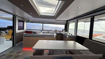 A imagem retrata o interior de um barco, com uma mesa e cadeiras, com uma grande janela ao fundo. A sala é bem iluminada e tem um design moderno. A atmosfera geral é de relaxamento e conforto.