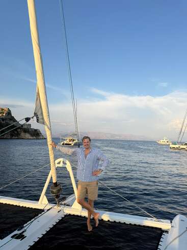 Um homem de camisa listrada e bermuda está descalço em um veleiro, cercado por um mar azul calmo, com outros barcos e penhascos rochosos nas proximidades.