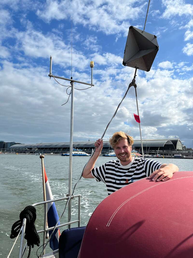 A imagem mostra um homem em pé em um barco, segurando uma corda, com um corpo de água e um céu nublado ao fundo, em uma paisagem urbana cênica.