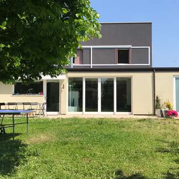 The image depicts the rear of a modern house with a patio and lawn, featuring a table and chairs, and a large tree in the foreground.