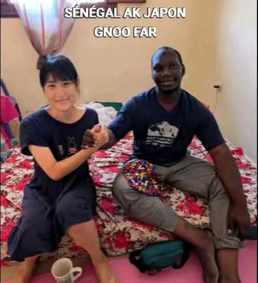 A Senegalese man and a Japanese woman sit on a bed, smiling and shaking hands, promoting cultural unity. The text above reads, "Sénégal ak Japon gnoo far."