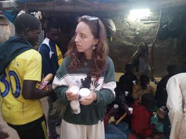 A woman stands holding cups and a water bottle, surrounded by people, some sitting and eating. The setting appears to be a modest, dimly-lit indoor area.