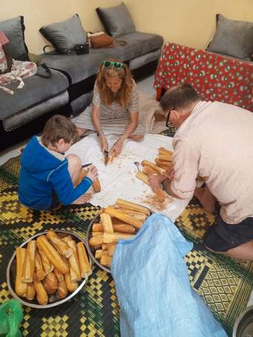 Three people sit on the floor preparing bread, surrounded by loaves and crumbs. They work together on mats in a cozy living room with couches and pillows.
