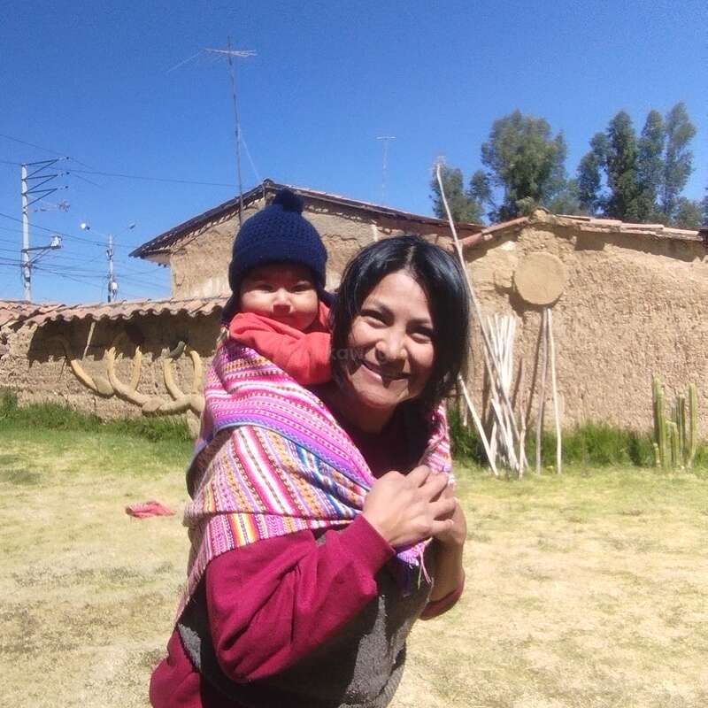 A woman smiles carrying a baby on her back with a colorful blanket. They stand outside on a sunny day, with blue sky and rustic buildings.