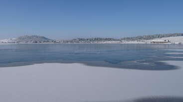 Eine ruhige Winterlandschaft mit einem teilweise zugefrorenen See, schneebedecktem Boden, fernen frostigen Hügeln, einem klaren blauen Himmel und einem einsamen Haus in Ufernähe.