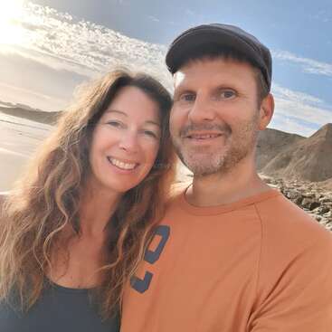 Um casal sorridente tira uma selfie em uma praia ensolarada. Há um fundo cênico com penhascos rochosos, costa arenosa e um lindo céu azul com nuvens.