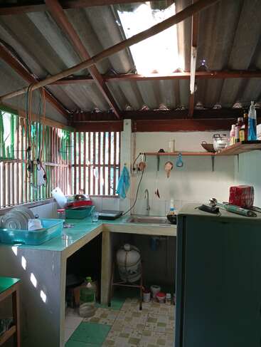 This image shows a small, rustic kitchen with green countertops, dishes, cleaning supplies, open wooden shelves, a water container, and sunlight streaming through a partially open roof.