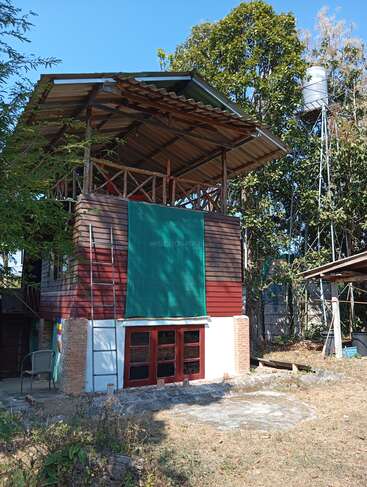 This image shows a two-story wooden house with a metal roof and open upper deck, surrounded by trees. A green shade covers part of the facade.