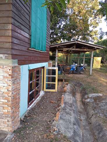 A rustic wooden house with an open yellow door, a nearby shed sheltering two motorbikes, surrounded by trees, gravel ground, and a concrete drainage ditch.