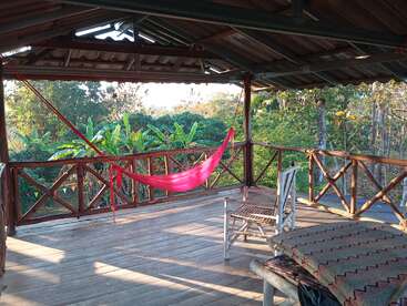 A peaceful wooden veranda overlooks lush green trees. A bright pink hammock hangs in the shade, next to a rustic rocking chair and cushioned bench. Serene retreat.
