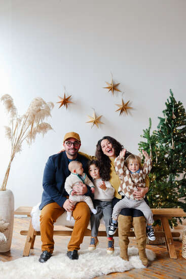 A joyful family of five sits on a wooden bench, surrounded by holiday decorations, a Christmas tree, star ornaments, and pampas grass, creating a festive atmosphere.
