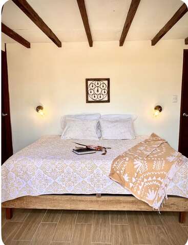 A cozy bedroom with a wooden bed, patterned bedding, two pillows, wall sconces, a decorative wall art, wooden beams, and a beige throw blanket.
