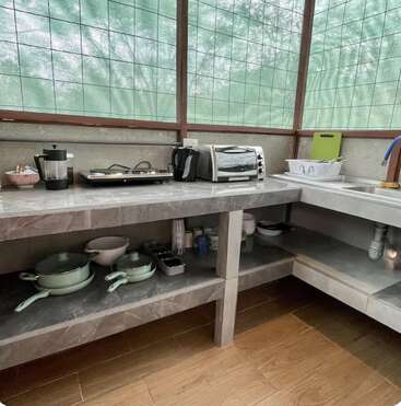 This kitchen features a marble countertop, toaster oven, kettle, French press, stove, dish rack, green pots, utensils, and open shelves with various bowls and glasses.