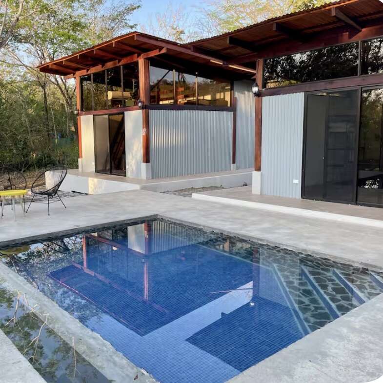 A modern house with large windows and corrugated metal walls sits in a forested area. In front, there’s a clean blue-tiled swimming pool and patio furniture.