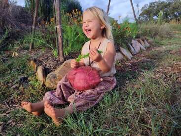 Uma jovem de cabelos loiros está sentada na grama, rindo, segurando uma grande batata-doce vermelha e uma planta verde. Ela usa uma camiseta regata bege e calça listrada.