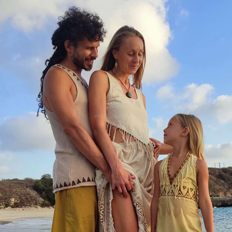 Uma família de três pessoas está reunida em uma praia sob um céu azul. Eles usam roupas de cores naturais e compartilham um momento caloroso, amoroso e gentil.