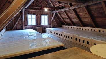 This cozy wooden attic room features multiple beds with plastic covers, rustic wooden walls, exposed beams, two windows, a bare bulb, and minimal furnishings.