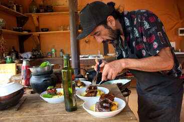 A chef carefully plates gourmet food in a rustic kitchen. Shelves with dishes and bottles line the background. Olive oil and fresh ingredients are visible.