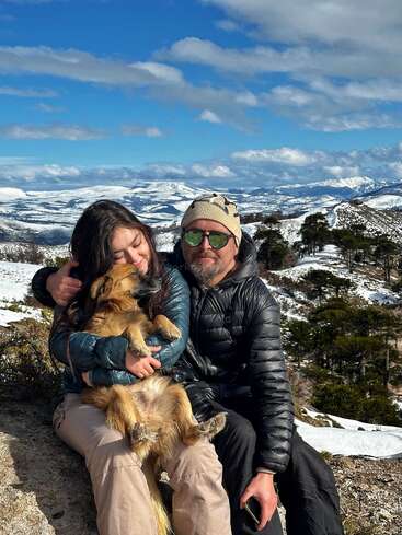 Ein Mann und eine Frau sitzen auf einem verschneiten Berg mit einem glücklichen Hund. Die Szenerie ist atemberaubend, voller Schnee, Bäume und blauem Himmel.