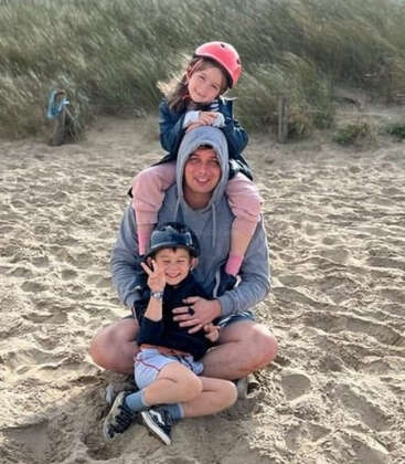 Ein Mann sitzt mit zwei lächelnden Kindern auf dem Sandboden, eines auf den Schultern, das andere vor ihm, beide tragen Helme, im Freien in der Nähe von grasbewachsenen Dünen.