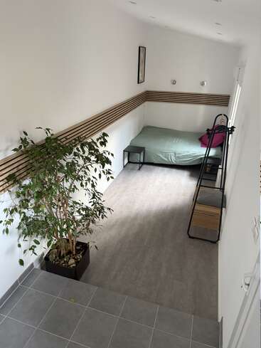 Dieses Bild zeigt ein minimalistisches Zimmer mit einem grünen Bett, zwei lila Kissen, einer Topfpflanze, kleinen Möbeln, weißen Wänden und modernen Holzpaneel-Akzenten.