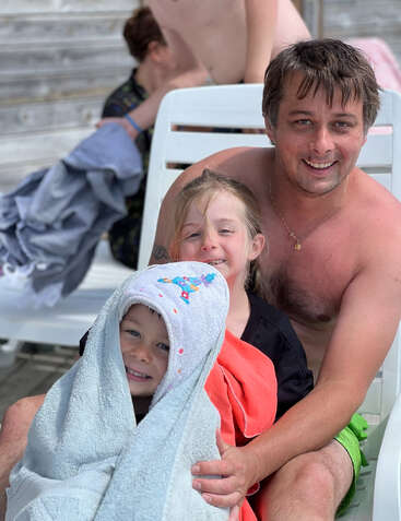 Ein lächelnder Mann sitzt auf einem Liegestuhl mit zwei glücklichen Kindern, eines davon in ein Handtuch gewickelt. Entspannende, sommerliche Atmosphäre mit einer Familie, die gemeinsam Zeit am Pool genießt.
