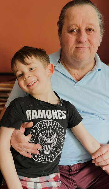 Um homem mais velho sorridente com uma camisa azul abraça um garoto feliz com uma camiseta dos "Ramones". Eles posam juntos contra um fundo marrom e quente.