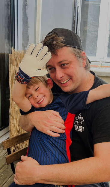 Um homem alegre e uma criança sorridente se abraçam. A criança veste uma camisa esportiva azul e luva de goleiro, irradiando felicidade e calor em um momento sincero.