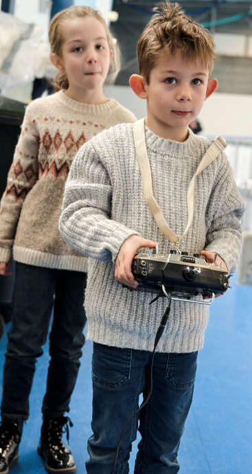 Um menino com um suéter cinza segura um dispositivo de controle remoto, enquanto uma menina fica atrás dele. As duas crianças parecem concentradas e intrigadas em um ambiente fechado.