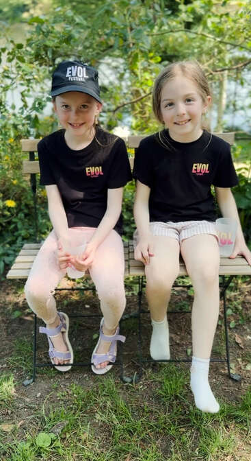 Duas garotas sorridentes sentadas em um banco de madeira ao ar livre, vestindo camisetas pretas combinando com o "EVOL Festival". Uma delas tem um boné, ambas segurando xícaras, cercadas por vegetação.