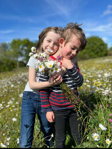 Duas crianças estão em um campo ensolarado de flores silvestres. A menina abraça o menino, ambos sorrindo, segurando um buquê de margaridas, desfrutando de um belo dia de primavera.