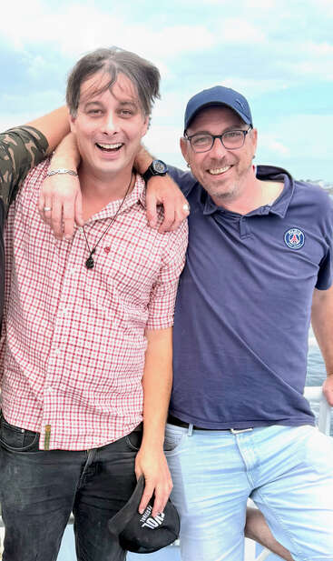 Dois homens estão sorrindo e posando juntos ao ar livre, um vestindo uma camisa xadrez e segurando um boné, o outro vestindo uma camisa polo azul e um boné.