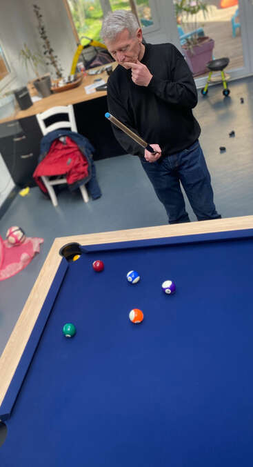 Um homem de suéter preto examina atentamente uma mesa de bilhar, com o taco na mão. Bolas coloridas estão espalhadas. O ambiente é casual, desorganizado e descontraído.
