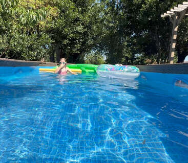 Uma criança usando óculos de proteção relaxa em uma piscina azul brilhante cercada por árvores. Boias coloridas e brinquedos de piscina flutuam nas proximidades, criando uma atmosfera ensolarada e tranquila.
