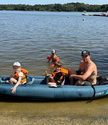 Um homem e duas crianças com coletes salva-vidas sentados em um barco inflável na praia, enquanto outra criança brinca na água em um dia ensolarado.