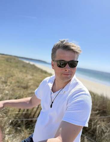 A man wearing sunglasses and a white shirt enjoys a sunny day by the beach, with blue sky, grassy dunes, and ocean visible in the background.