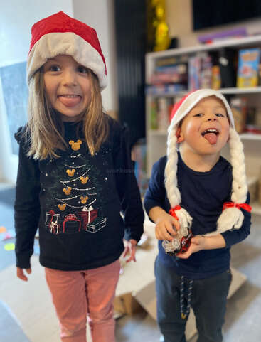 Zwei Kinder mit festlichen Weihnachtsmannmützen lächeln verspielt und strecken ihre Zungen heraus. Sie stehen in einem Haus, umgeben von Spielzeug und Geschenken, und strahlen fröhliche Weihnachtsstimmung aus.