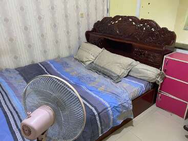 Esta imagen muestra un pequeño dormitorio con un cabecero de madera tallada, sábanas con motivos azules, varias almohadas, un ventilador de pie rosa y una cómoda rosa.