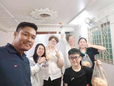 Un grupo de seis personas posan felices en el interior, sosteniendo bolsas con pan. Sonríen y parecen alegres, probablemente celebrando o compartiendo comida juntos en una habitación luminosa.