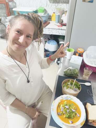Una mujer sonriente vestida de blanco está sentada a la mesa, haciendo el signo de la paz, rodeada de deliciosa comida, sopa, ensalada y pan, en una acogedora cocina.