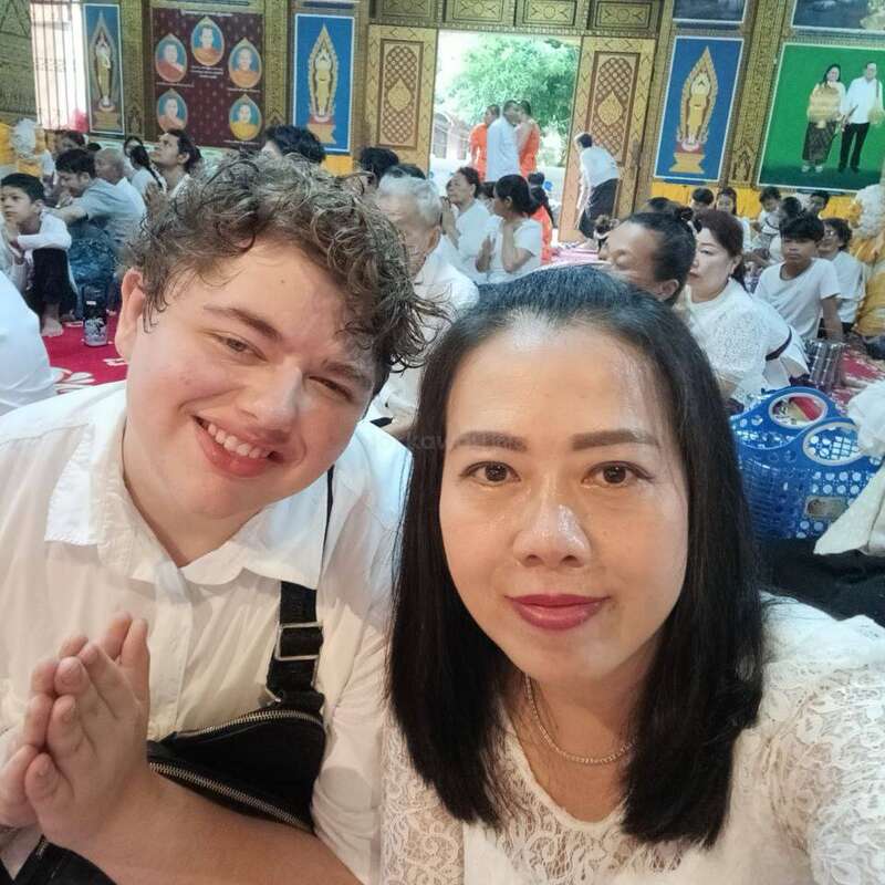 A smiling man and woman pose for a selfie inside a colorful, ornate temple. Many people dressed in white gather in the background, participating in a religious event.