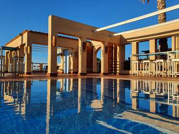 Das Bild zeigt eine ruhige Poolszene mit einem hellbraunen Gebäude mit überdachtem Terrassenbereich, umgeben von Palmen und einem klaren blauen Himmel.