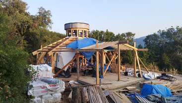 Una estructura redonda de madera parcialmente construida se alza en un claro del bosque. Hay materiales de construcción y lonas azules esparcidos mientras los trabajadores montan el edificio de estructura abierta.
