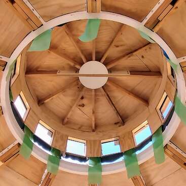 Esta imagen muestra el interior de una cúpula de madera o techo de yurta. La luz del sol entra por ventanas rectangulares, y dentro cuelgan tiras de tela verde que crean un ambiente luminoso.